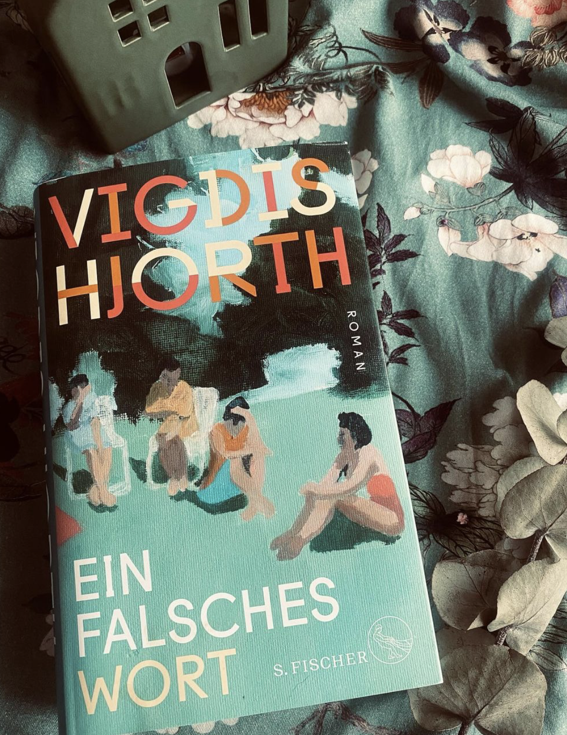Ein Buch auf einem bedruckten Tuch. Das Cover zeigt mehrere Personen, die an einem Strand sitzen. Ein falsches Wort